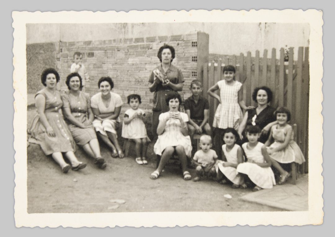 Ceferina García García’s daughters having an afternoon snack (merienda) with one of Ceferina’s aunts, the neighbours, and their children in front of the house gate. The aunt is holding a tambourine (pandereta); while she played, the children sang along. At that time, Ceferina and her husband were living in Germany, while their daughters remained in Spain with the aunt. Vega-Gijón, 1962 © Cristina Fernández-Moser